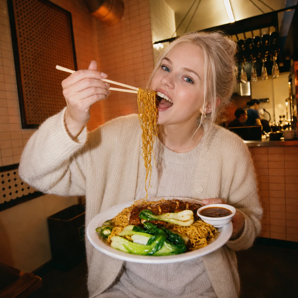 Smiling diner in a cream knit sweater holding up a plate of stir-fried noodles with bok choy and dipping sauce, lifting a bite to her mouth with chopsticks in a warm-lit noodle bar.