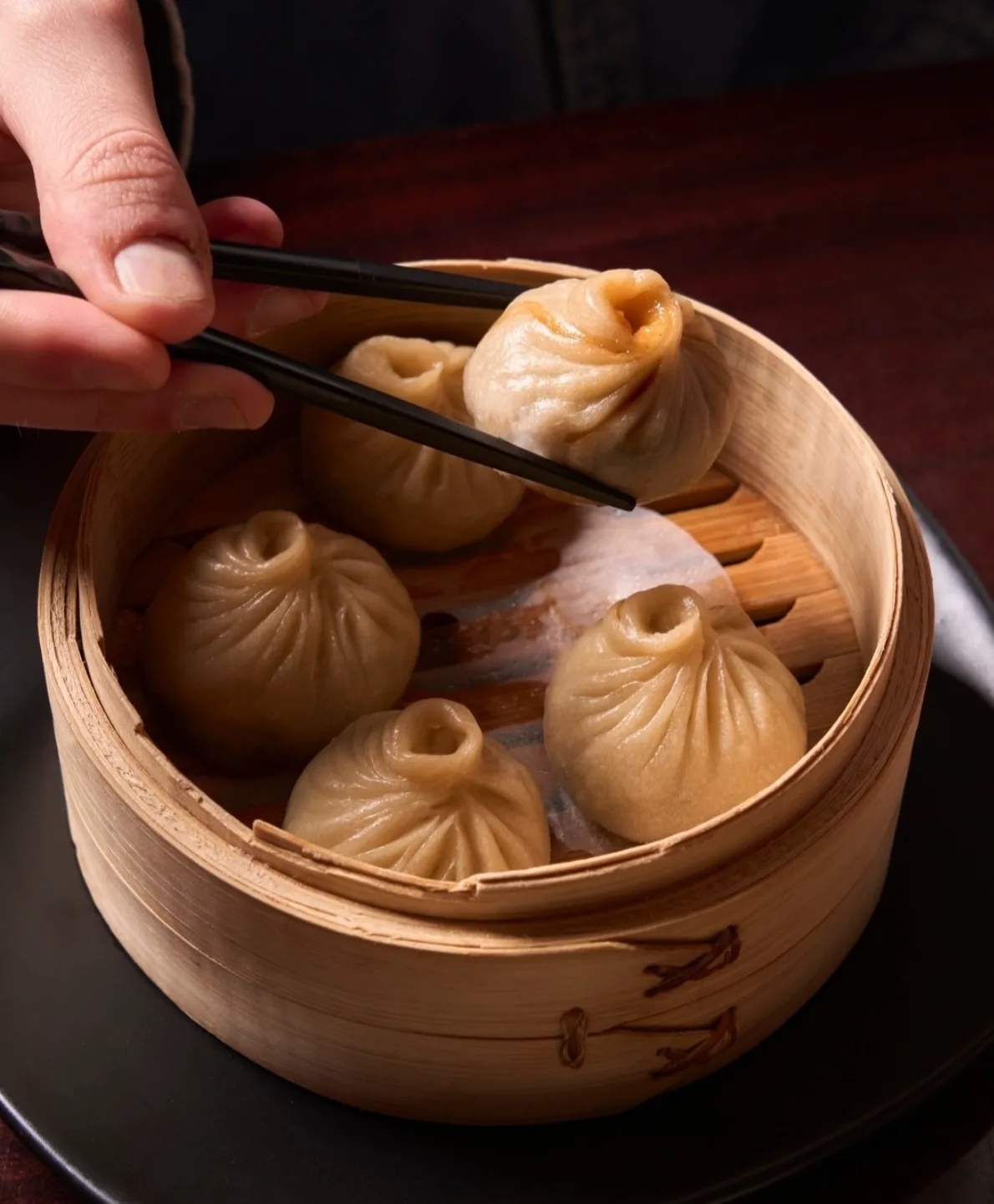 Bamboo steamer of xiao long bao soup dumplings with a hand lifting one out with chopsticks.