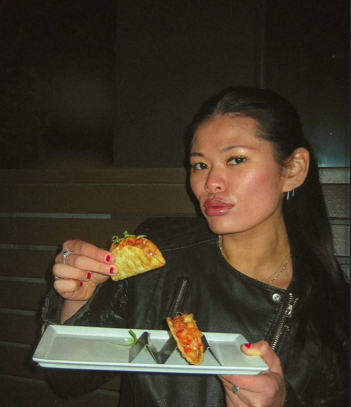 Woman in a leather jacket holding a small taco and a tray of more tacos, looking directly at the camera in low restaurant lighting.