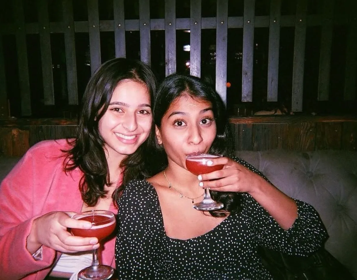 Two friends smiling and holding pink cocktails in a dim restaurant booth.