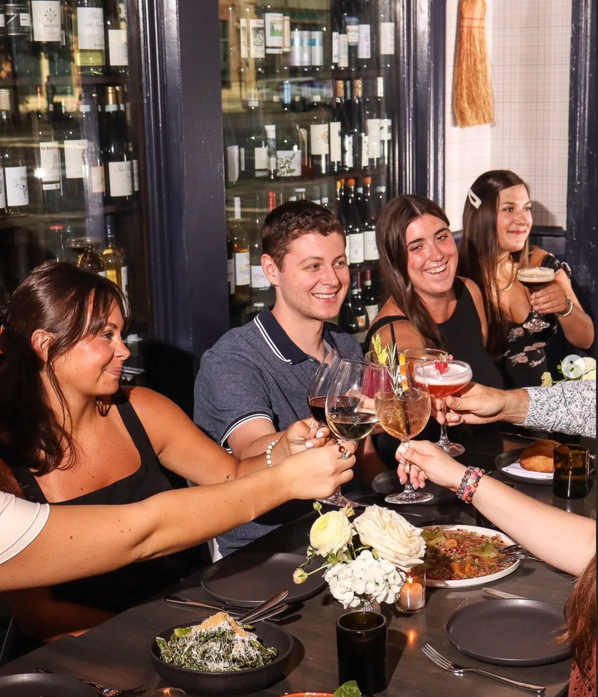 Group of friends laughing and clinking glasses of wine and cocktails at a bar lined with bottles.